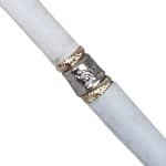 A close-up of a white cylindrical product, likely a candlestick or decorative tube, featuring a metallic silver and gold band with an embossed floral design.