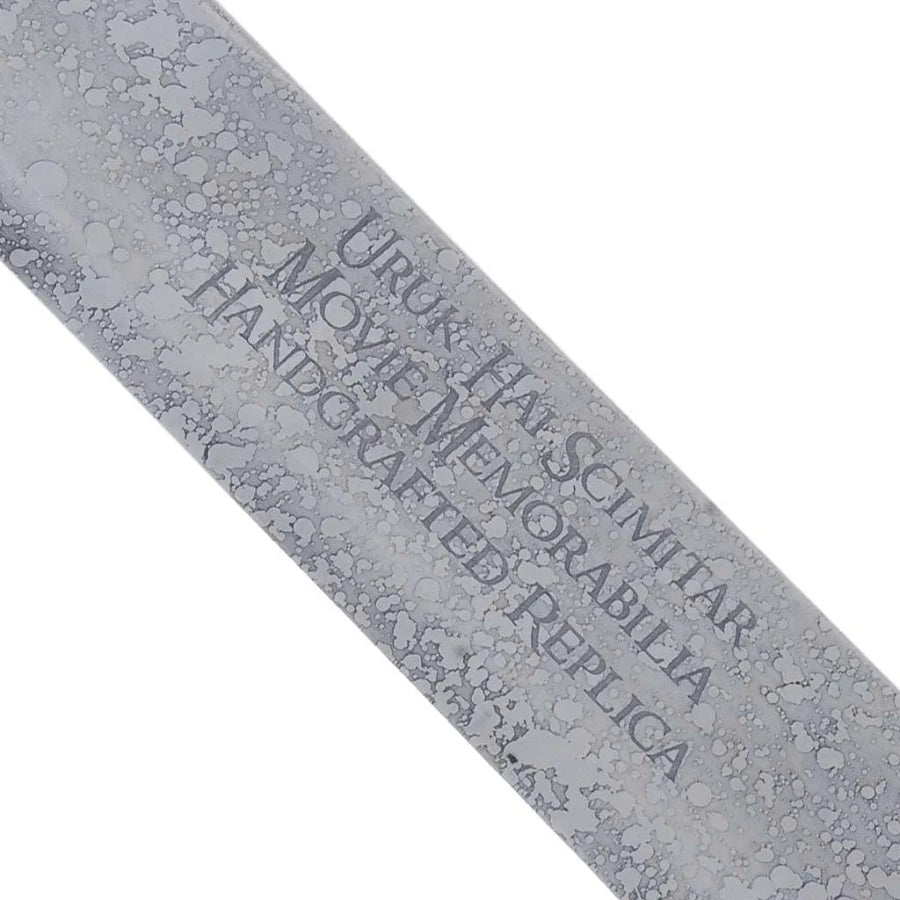 Close-up of a metallic silver replica sword blade with a textured, hammered finish and engraved text reading 'URUKHAI SCIMTAR MOVIE MEMORABILIA HANDCRAFTED REPLICA'.